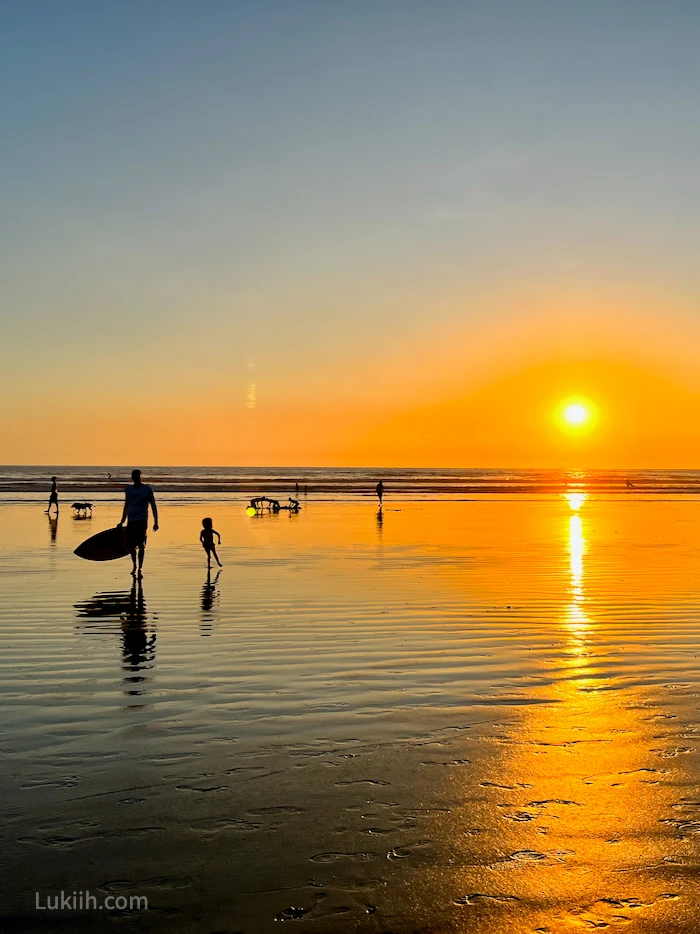 Silhouettes of a man carrying a surfboard against a sunset dipping into the ocean.