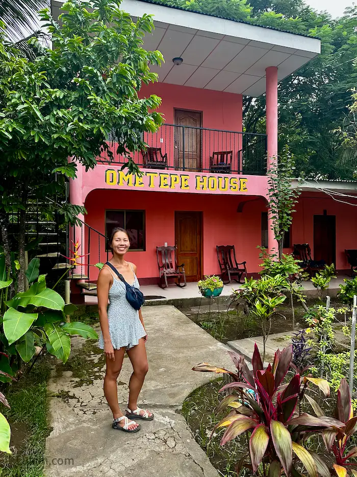 A woman standing in front of a pink building that says Ometepe House.