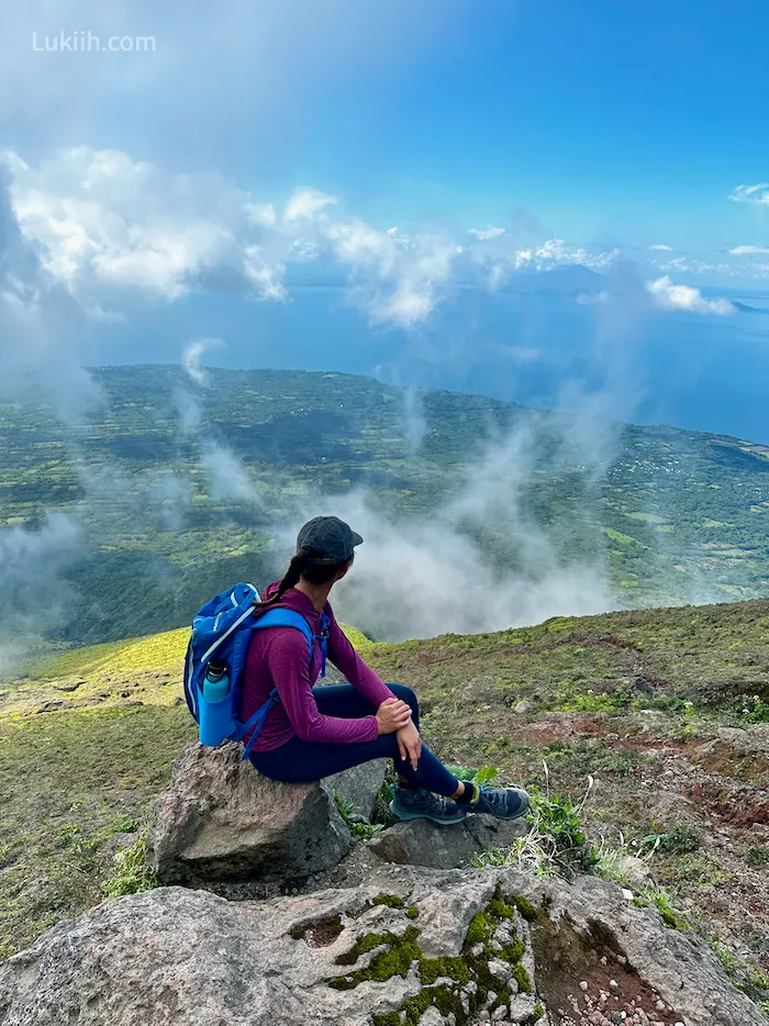 A woman sitting down on a mountain looking at a view with green land surrounded by ocean.