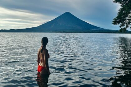 A woman partially submerged in lake water while looking at a volcano in the background.