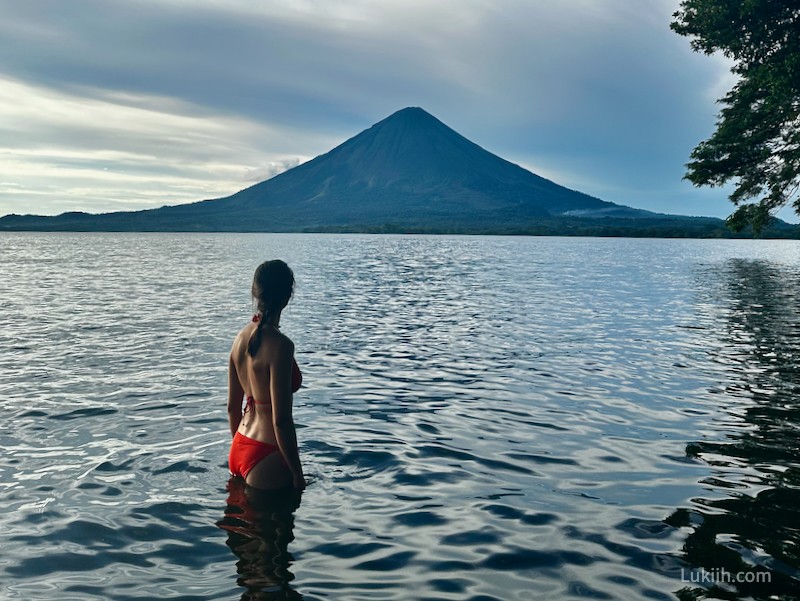 A woman partially submerged in lake water while looking at a volcano in the background.