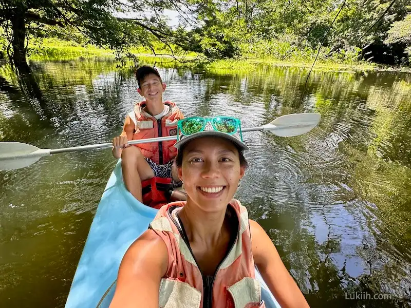 Two people kayaking over water surrounded by lush trees.