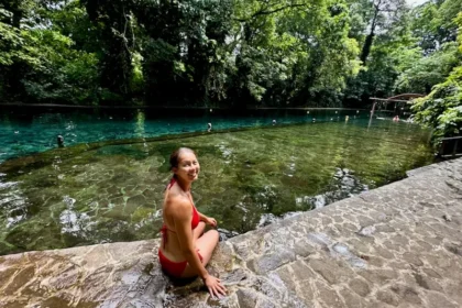 A woman sitting at the edge of a clear pool in a tropical setting.