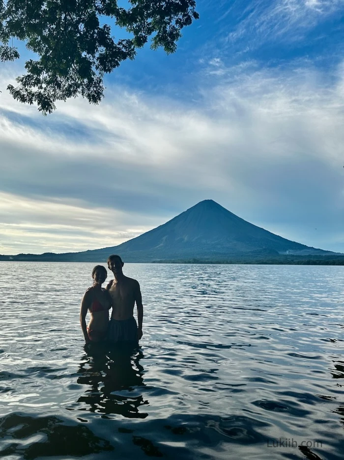 Two people submerged in lake water while looking at a volcano in the background.