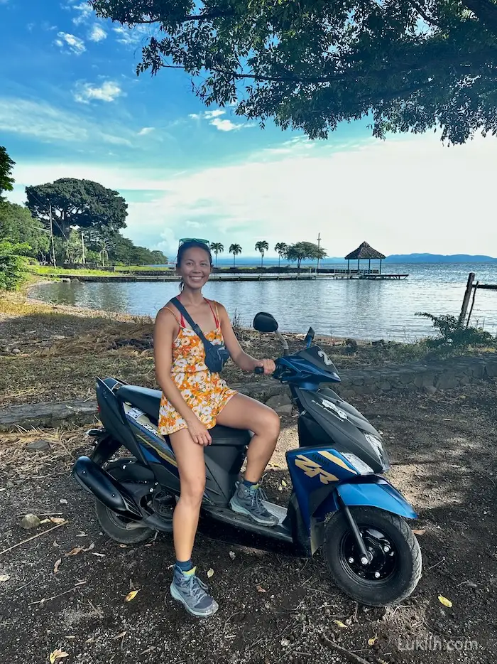 A woman sitting on a scooter with a lake and palm trees in the background.