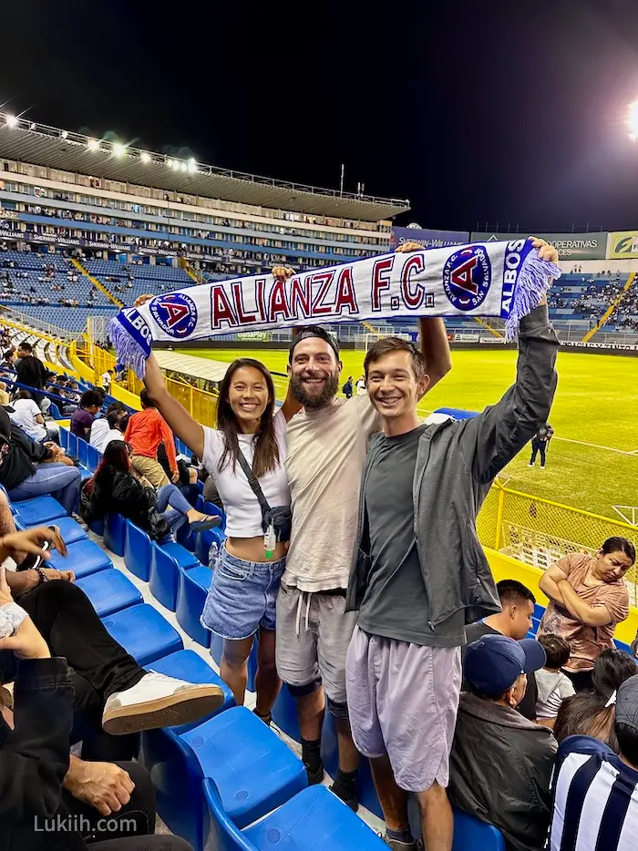 Three people holding a sign that says Alianza FC at a stadium.