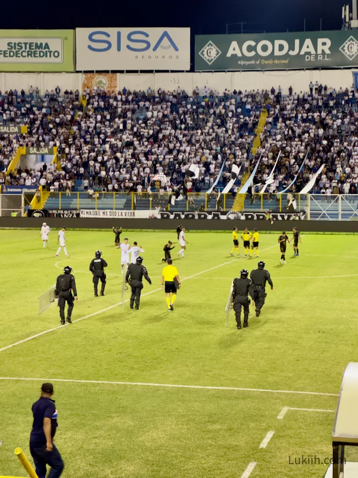 A referee walking on a soccer field protected by police in riot gear.