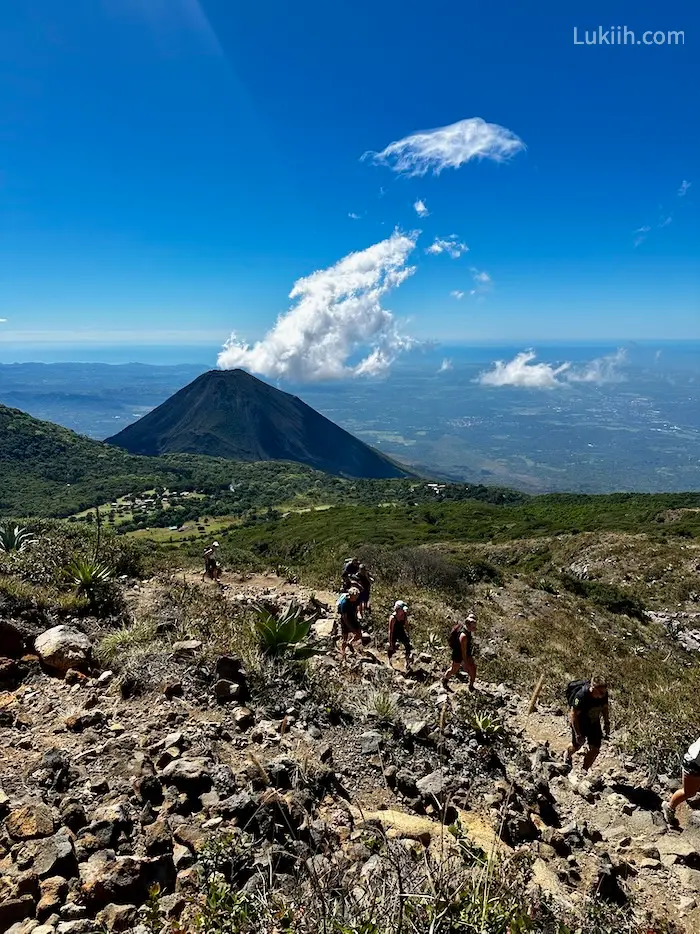 Hikers walking on a dirt trail with a view of a volcano in the background.