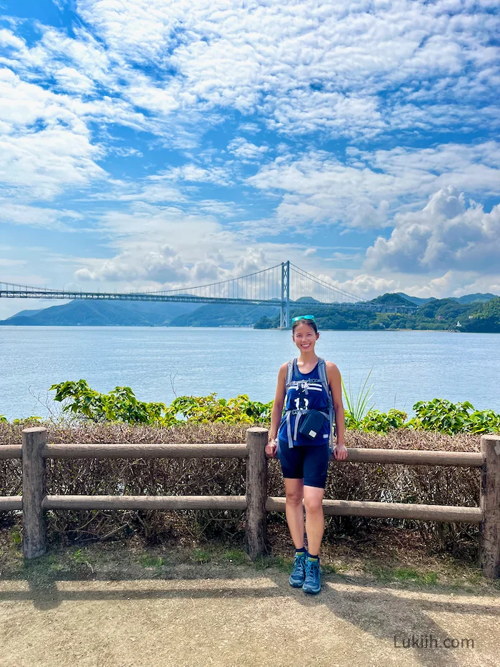 A woman in a biking outfit standing next to the ocean with a bridge in the background.