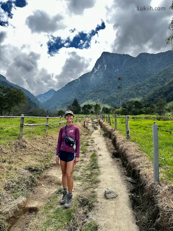 A hiker standing on a dirt trail surrounded by lush grass and a big mountain peak in the background.