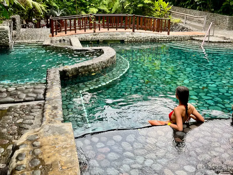 A woman leaning in a quiet pool with a bridge and lush trees in the background.