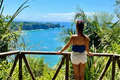 A woman standing at a viewpoint overlooking a blue ocean and lush rainforest.