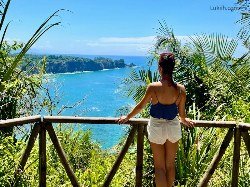 A woman standing at a viewpoint overlooking a blue ocean and lush rainforest.