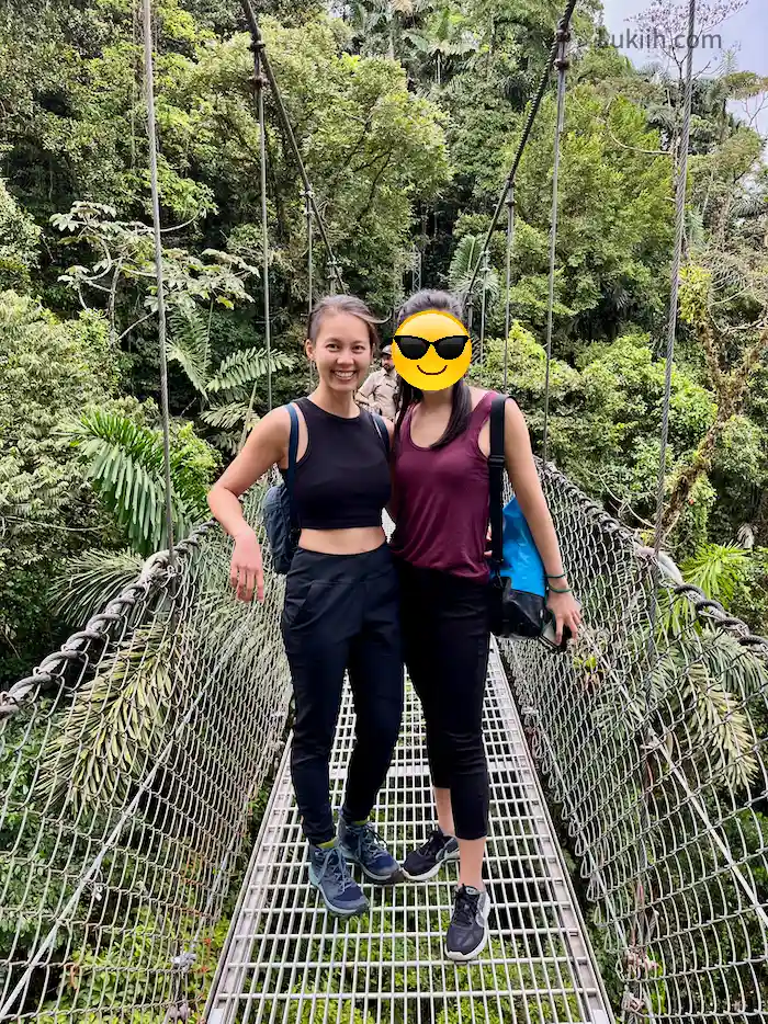 Two people standing on a suspended bridge surrounded by rainforest.