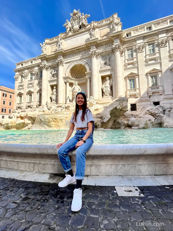 A woman sitting in front of a white, marble fountain.