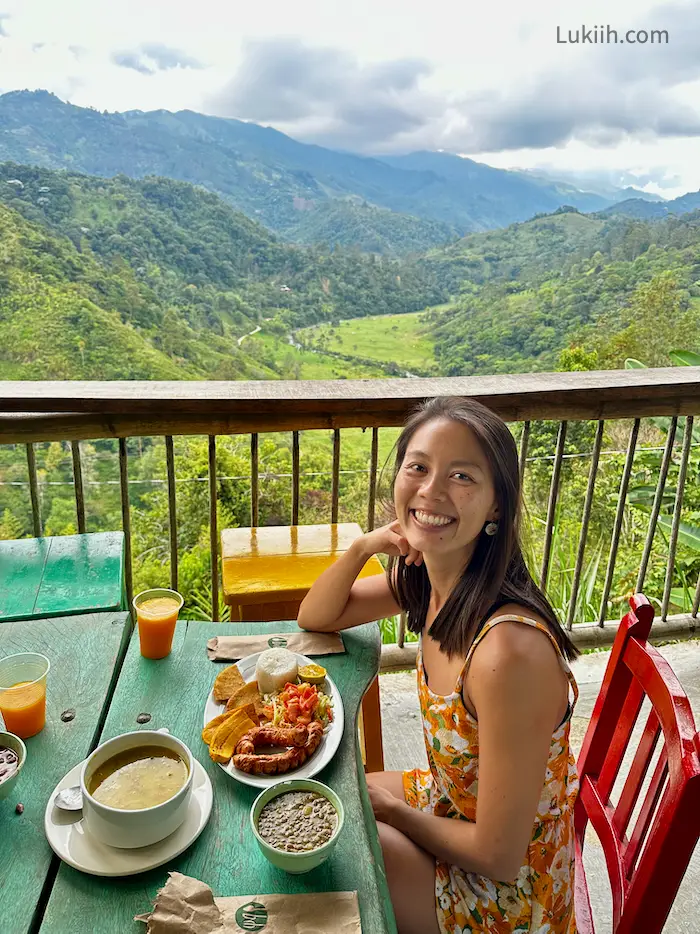 A woman eating a plate with rice, plaintains, sausage, and soup with a view of a lush valley.