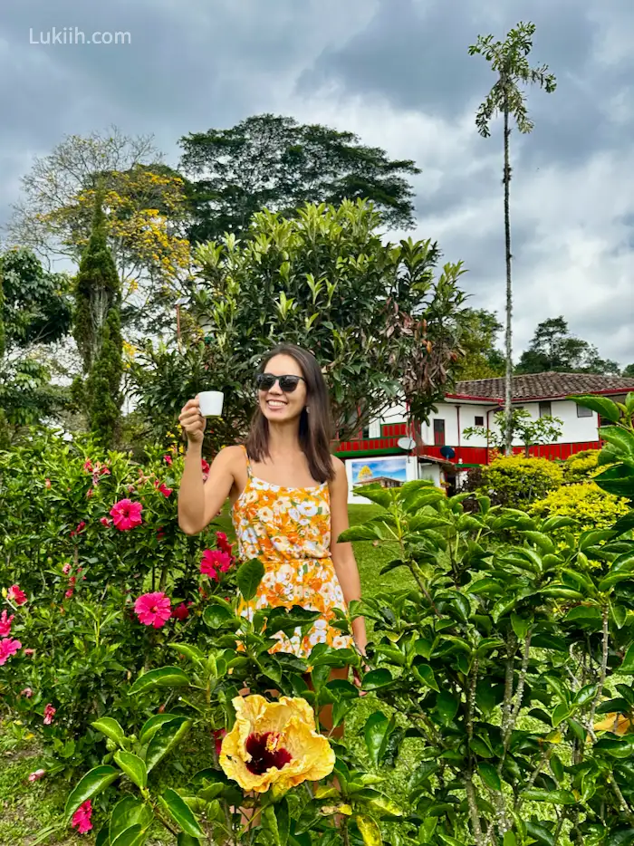 A woman holding a cup of coffee in a lush area with flowers.