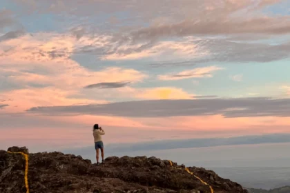 A woman standing on a high rock taking a photo of a sunset.