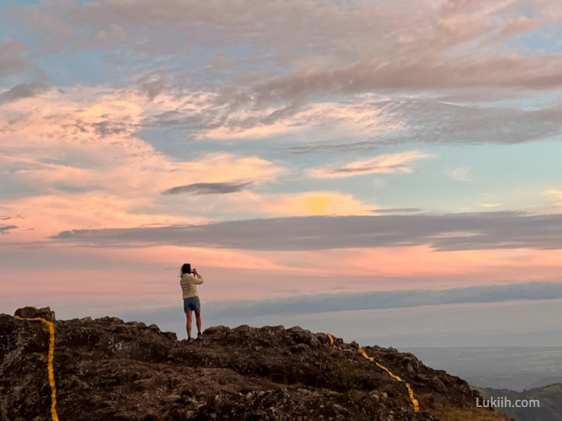 A woman standing on a high rock taking a photo of a sunset.