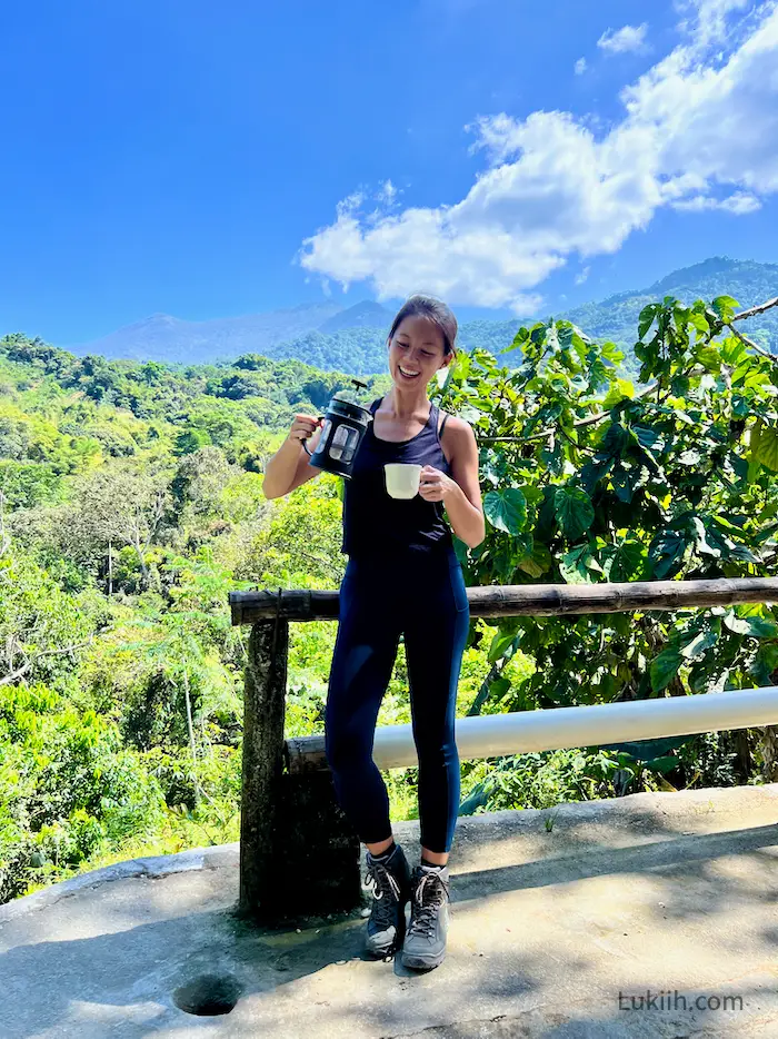 A woman pouring coffee against a lush mountain background.