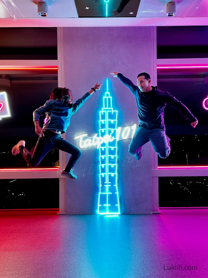 Two people jumping and posing for a photo against a "Taipei 101" background.