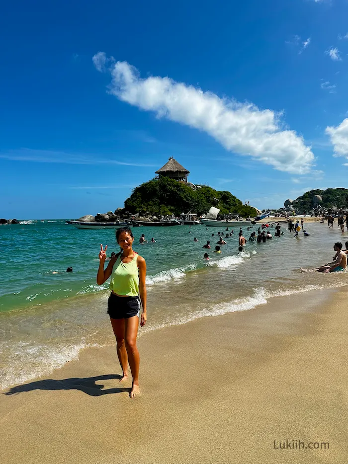 A woman standing on a sandy beach with a hut and many people in the background.