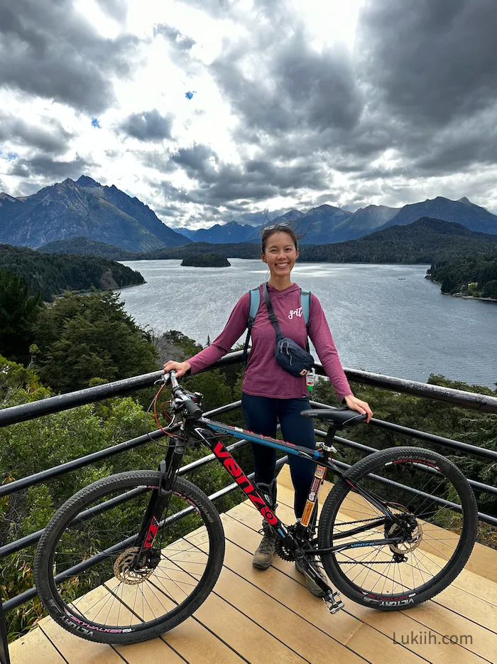 A woman with a mountain bike on a lookout point with lakes and mountains in the background.