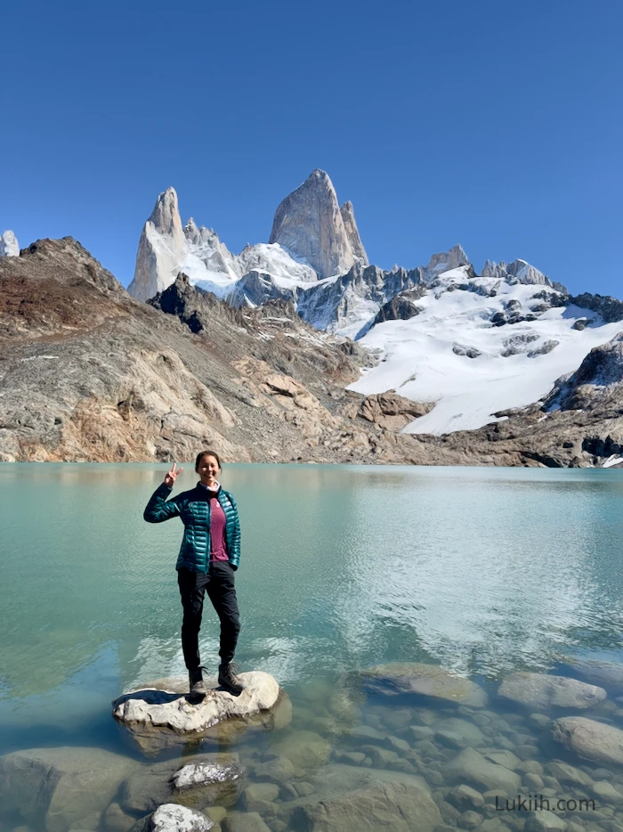 A woman standing on a rock on a lake with a distinct mountain peak in the background.