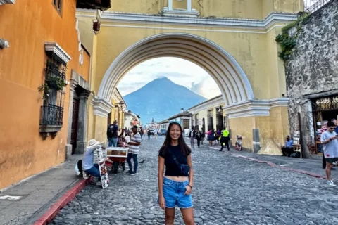 A woman standing in front of a cobblestone city with a volcano in the background.